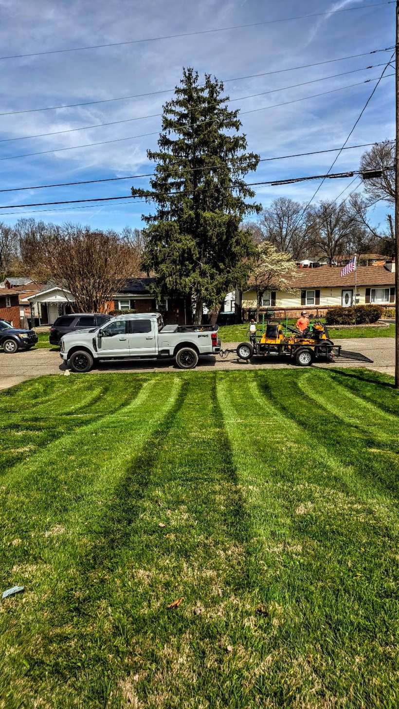 A front lawn cut in clean alternating stripes running down to the kerb, with a pickup truck and equipment trailer parked at the top of the drive.