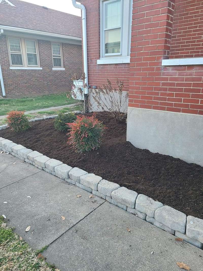 A long planting bed edged in pale stone blocks alongside a concrete walkway, freshly mulched, with a brick house behind.