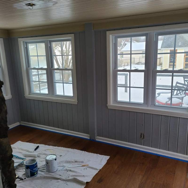 A room mid-decoration with a drop cloth over the floor, paint cans and a roller tray, taped trim and bare grey panelling.